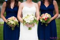 Bridesmaids in navy dresses hold coral, white, peach, and green bouquets, while the bride carries white, ivory, and green flowers. By Floressence.